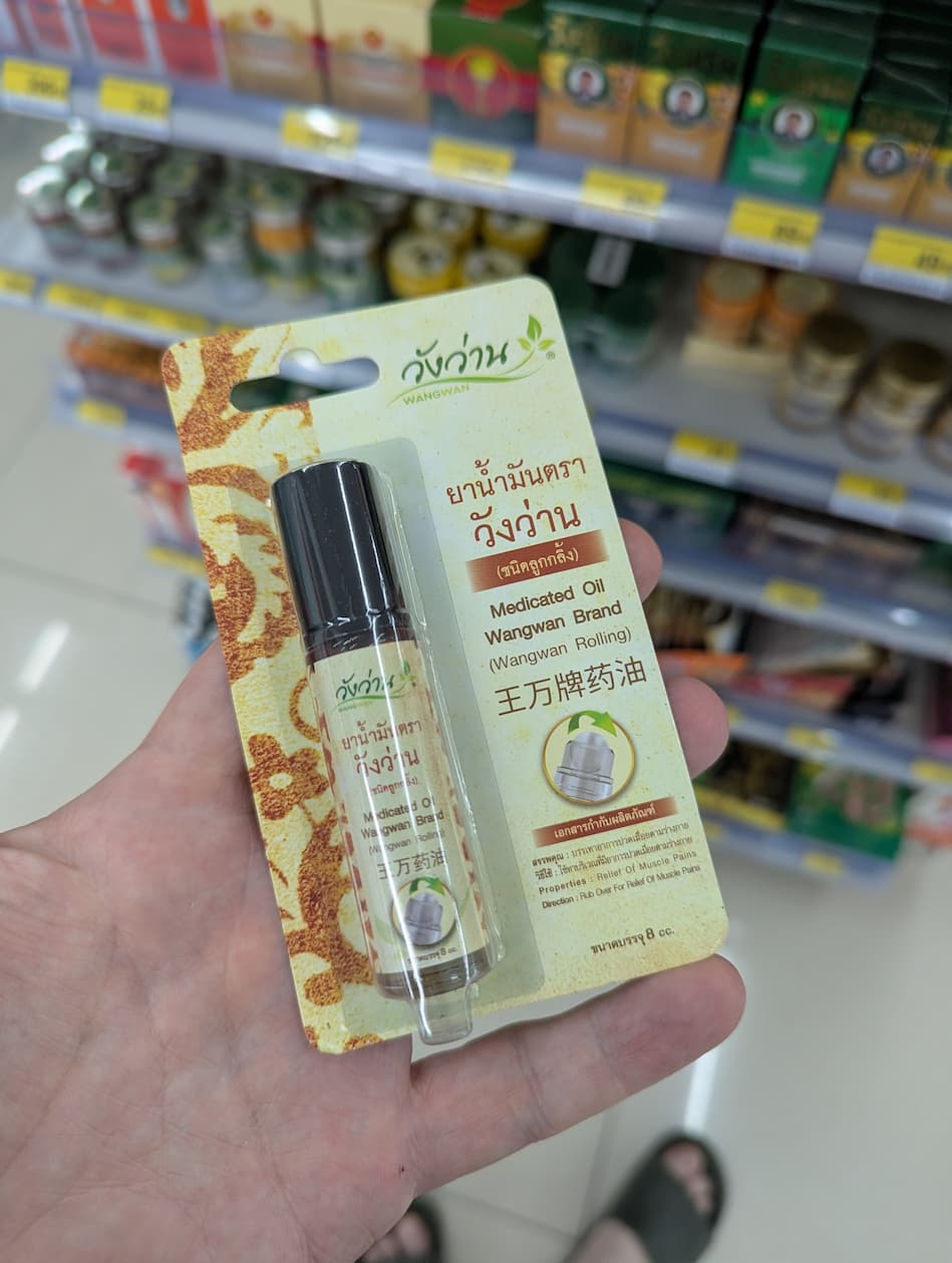 Hand holding a Wangwan Medicated Oil roll-on pack in a Thailand 7-Eleven pharmacy aisle