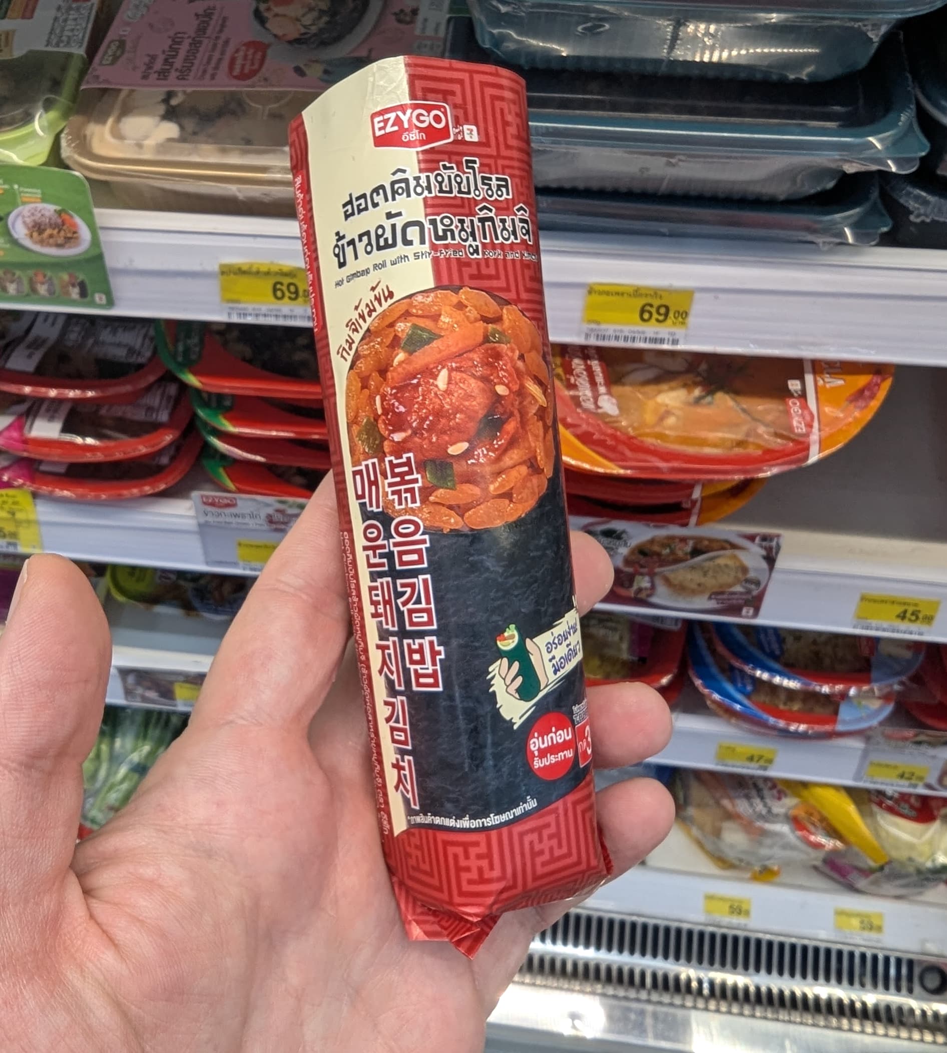A person holding a packaged Ezygo Hot Gimbap Roll with stir-fried pork and kimchi from 7-Eleven Thailand.