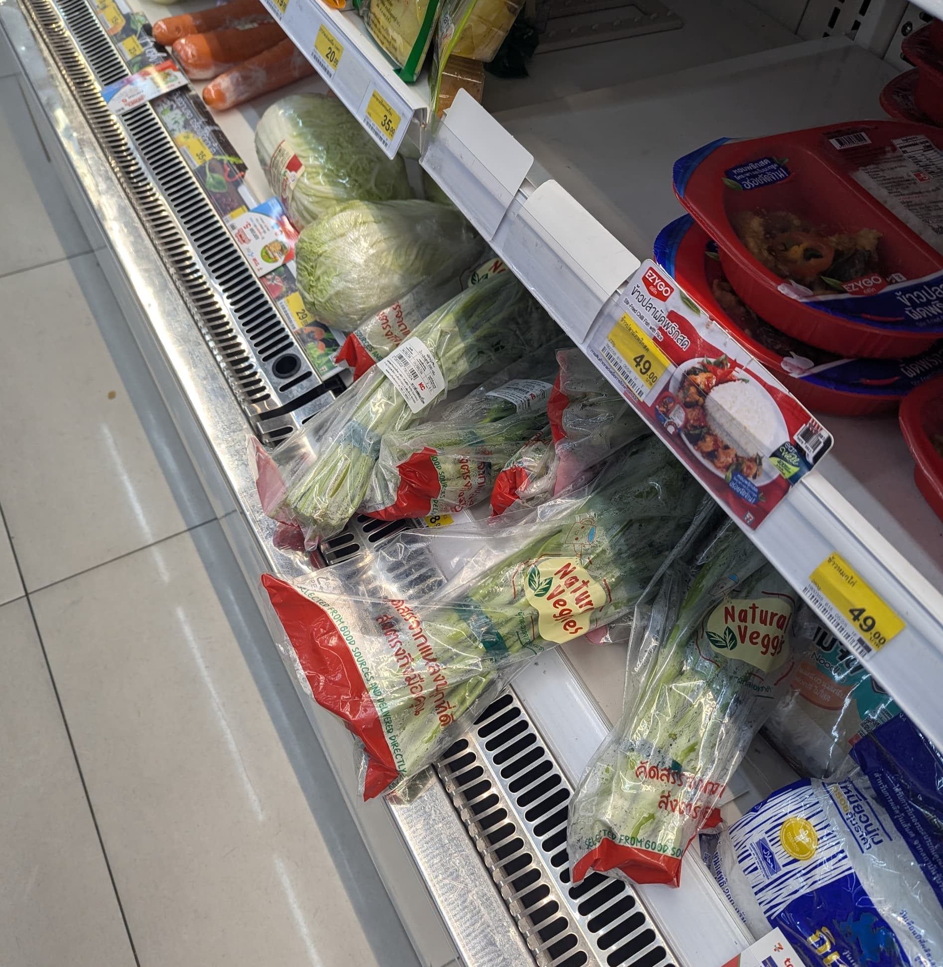 A refrigerated shelf at 7-Eleven Thailand showing bags of fresh 'Natural Veggies' including morning glory and cabbage.
