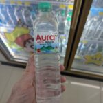 Soffell Mosquito Repellent Lotion: The Essential 7-Eleven Defense Against Thai Mosquitoes A hand holding a transparent plastic bottle of Aura 100% Natural Mineral Water with a green cap inside a 7-Eleven in Thailand.