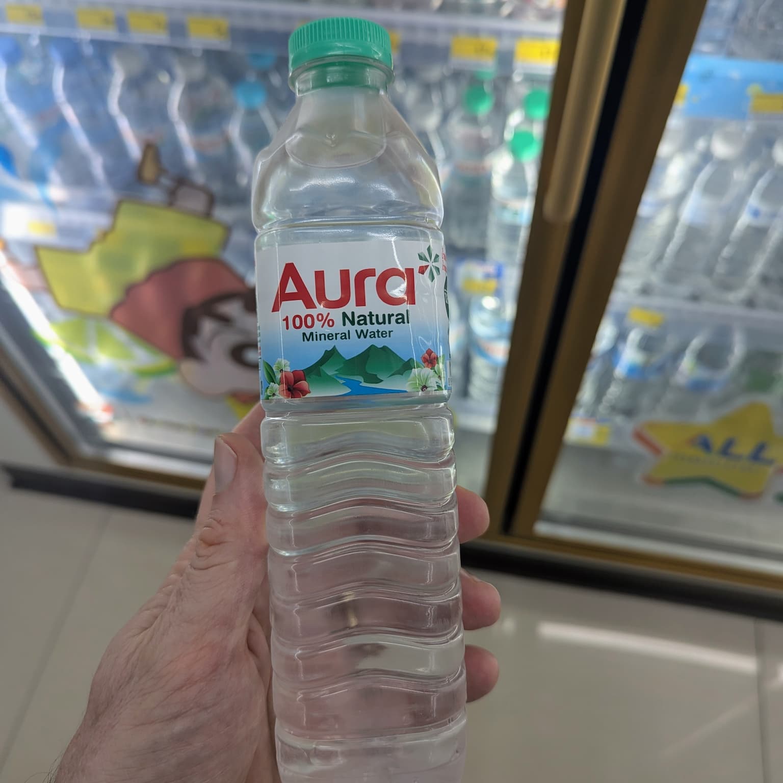 A hand holding a transparent plastic bottle of Aura 100% Natural Mineral Water with a green cap inside a 7-Eleven in Thailand.
