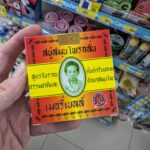 A hand holding a square red, yellow, and green box of Madame Heng Original Herbal Soap inside a Thai store.