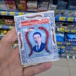 A hand holding a white sachet of Takabb Anti-Cough Pills with centipede illustrations inside a 7-Eleven store in Thailand.