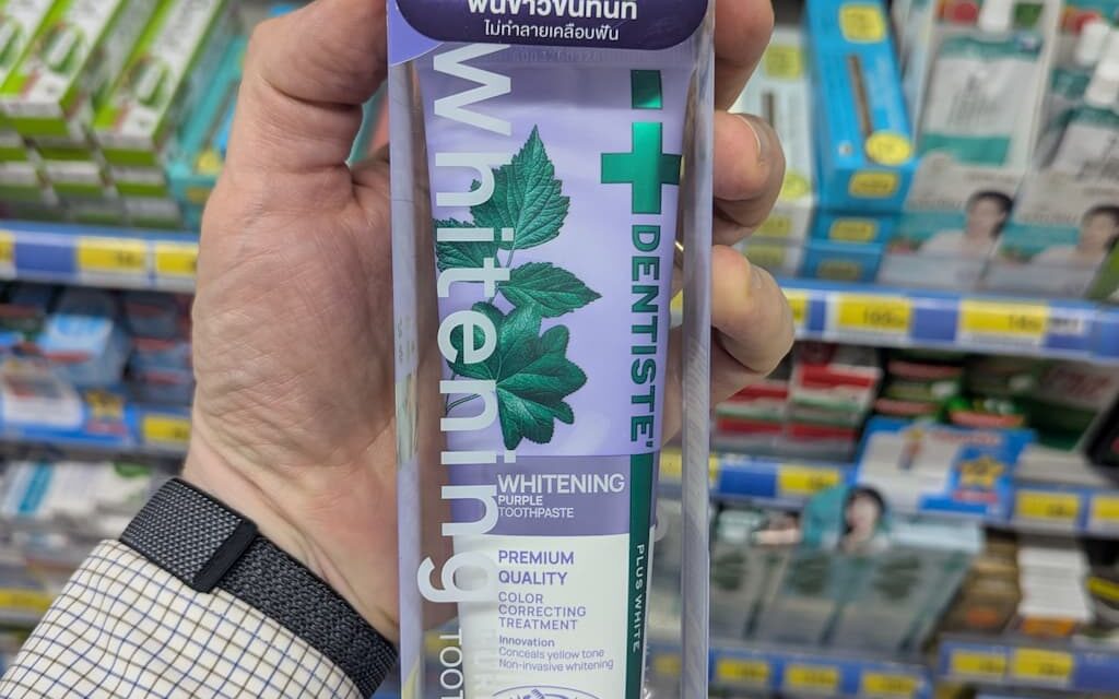 A hand holding a box of Dentiste' Whitening Purple Toothpaste featuring Lisa from Blackpink inside a store in Thailand.