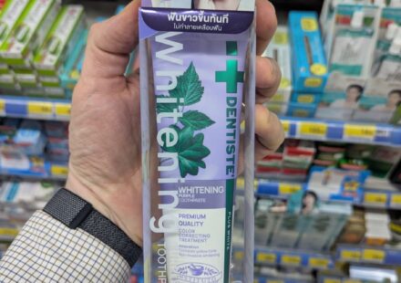 A hand holding a box of Dentiste' Whitening Purple Toothpaste featuring Lisa from Blackpink inside a store in Thailand.