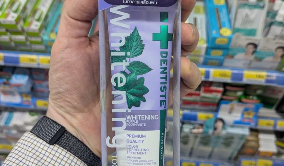 A hand holding a box of Dentiste' Whitening Purple Toothpaste featuring Lisa from Blackpink inside a store in Thailand.