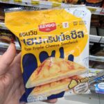 A hand holding a yellow and blue plastic pouch packaging of an Ezygo Ham Triple Cheese Sandwich in front of store shelves in Thailand.