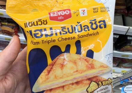 A hand holding a yellow and blue plastic pouch packaging of an Ezygo Ham Triple Cheese Sandwich in front of store shelves in Thailand.