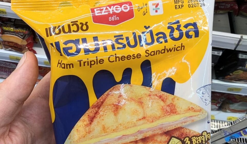 Ezygo Ham Triple Cheese Sandwich: The Ultimate Molten Melt Centerpiece A hand holding a yellow and blue plastic pouch packaging of an Ezygo Ham Triple Cheese Sandwich in front of store shelves in Thailand.