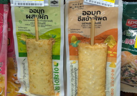Happy Muchy Korean Fishcake Sticks: A Trendy, Interactive Street Food Experience Green and orange vacuum-sealed packaging pouches of Happy Muchy Brand Korean Style Fishcake Sticks on a chilled 7-Eleven store shelf in Thailand.