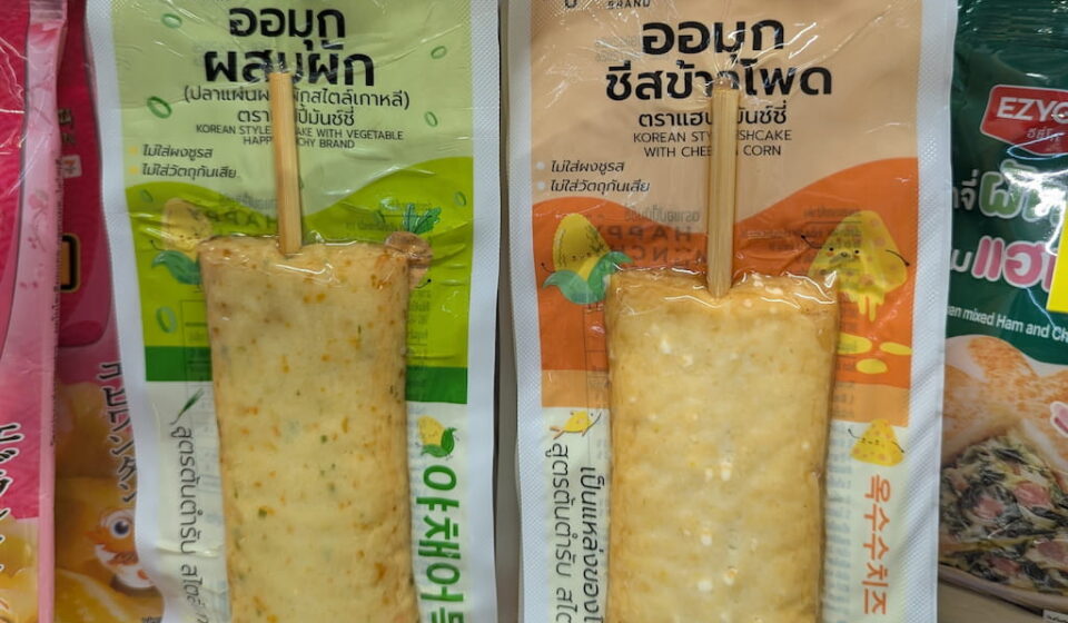 Green and orange vacuum-sealed packaging pouches of Happy Muchy Brand Korean Style Fishcake Sticks on a chilled 7-Eleven store shelf in Thailand.