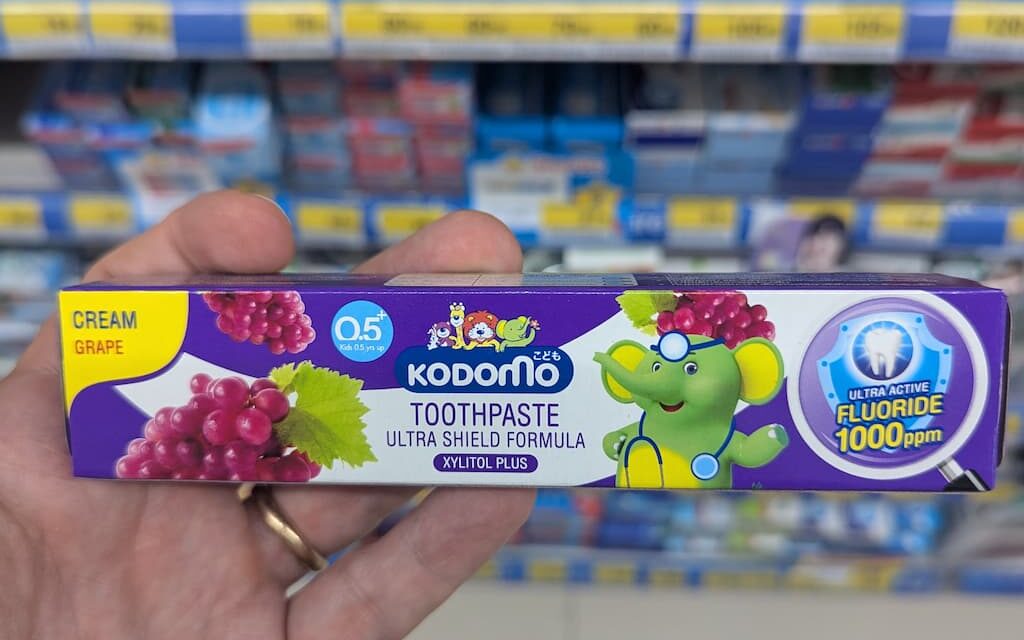 A hand holding a purple box of Kodomo Toothpaste Ultra Shield Formula in Cream Grape flavor inside a store in Thailand.