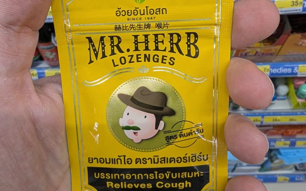 A hand holding a small yellow metallic pouch of Mr. Herb Lozenges for coughing relief inside a 7-Eleven store in Thailand.