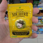 A hand holding a small yellow metallic pouch of Mr. Herb Lozenges for coughing relief inside a 7-Eleven store in Thailand.
