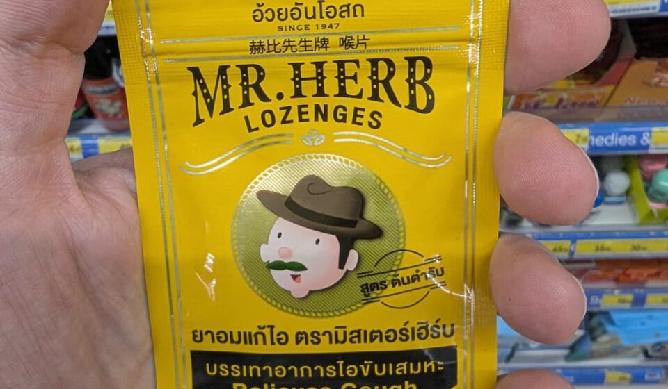 Mr. Herb Lozenges: The Authentic Herbal Rescue for a Scratchy Throat A hand holding a small yellow metallic pouch of Mr. Herb Lozenges for coughing relief inside a 7-Eleven store in Thailand.