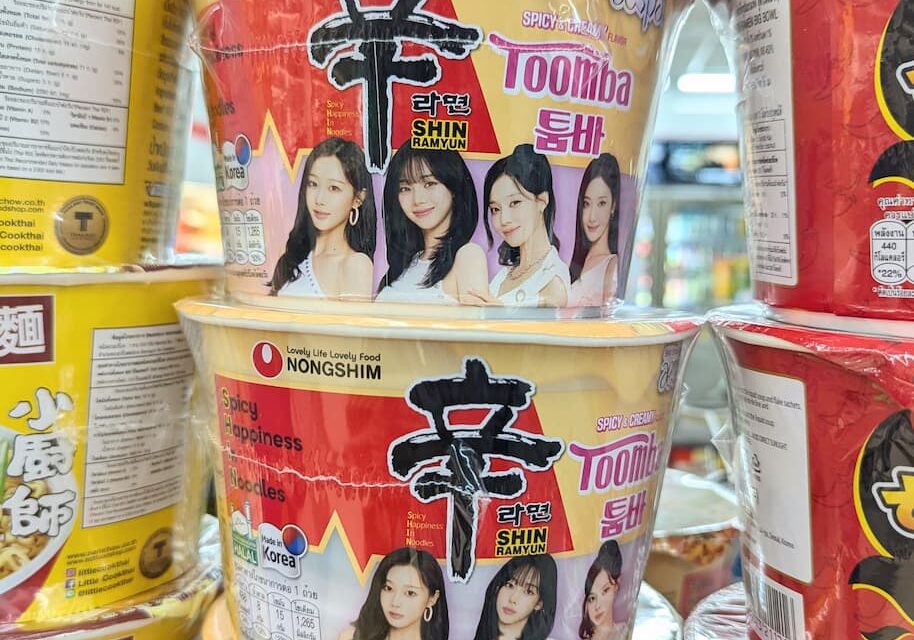 Yellow and cream insulated instant noodle bowls of Nongshim Shin Ramyun Toomba featuring K-pop group Aespa on a store shelf in Thailand.