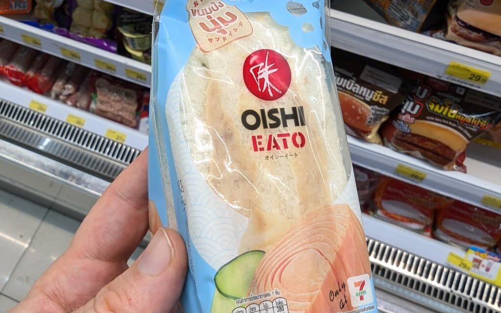 A hand holding a light blue plastic wedge packaging pouch of an Oishi Eato Tuna Salad Sandwich in front of store refrigerator shelves in Thailand.