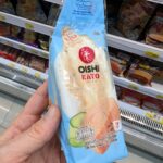 A hand holding a light blue plastic wedge packaging pouch of an Oishi Eato Tuna Salad Sandwich in front of store refrigerator shelves in Thailand.