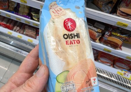 Oishi Eato Tuna Salad Sandwich: A Fresh and Soft Japanese-Style Cold Fix A hand holding a light blue plastic wedge packaging pouch of an Oishi Eato Tuna Salad Sandwich in front of store refrigerator shelves in Thailand.