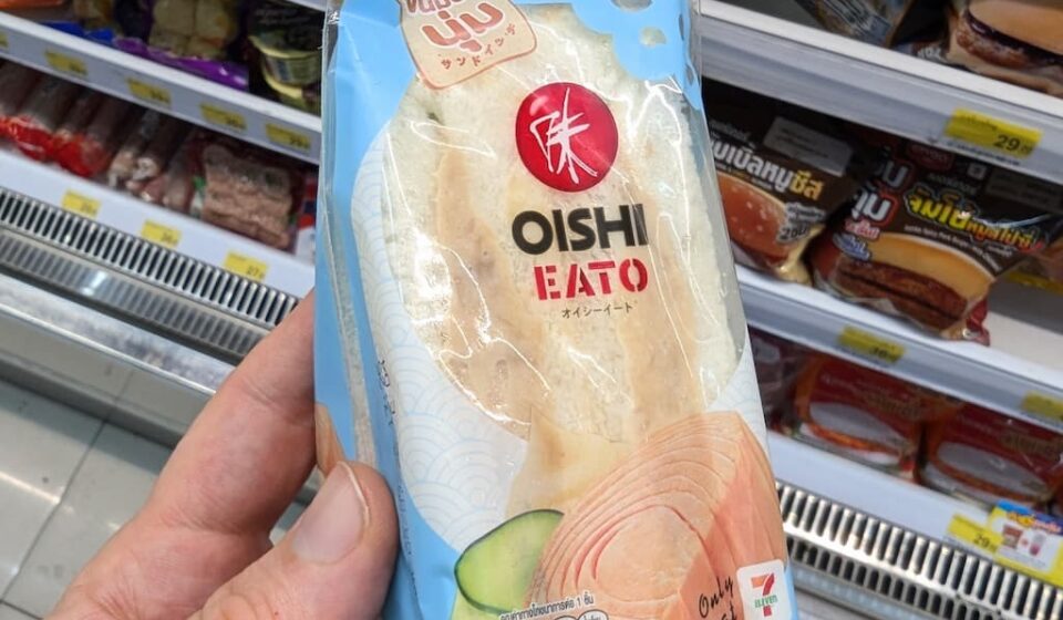 Oishi Eato Tuna Salad Sandwich: A Fresh and Soft Japanese-Style Cold Fix A hand holding a light blue plastic wedge packaging pouch of an Oishi Eato Tuna Salad Sandwich in front of store refrigerator shelves in Thailand.