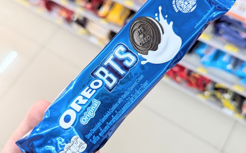 A hand holding a blue metallic plastic tube packaging of limited edition Oreo Original BTS cookies inside a store in Thailand.
