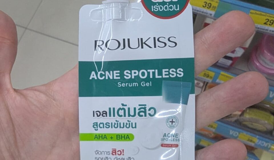 Rojukiss Acne Spotless Serum Gel: High-Quality Korean Skincare in a Pocket-Sized Sachet A hand holding a white and green sachet of Rojukiss Acne Spotless Serum Gel with a screw cap inside a store in Thailand.