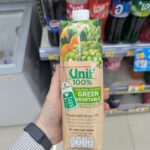 A hand holding a beige paper carton brik of Unif 100% Mixed Vegetable & Fruit Juice with Green Vegetable inside a 7-Eleven store in Thailand.