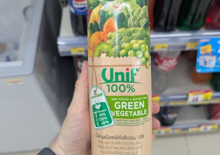 Unif 100% Green Vegetable Juice: A Cloying Mosaic of Industrial Leftovers A hand holding a beige paper carton brik of Unif 100% Mixed Vegetable & Fruit Juice with Green Vegetable inside a 7-Eleven store in Thailand.