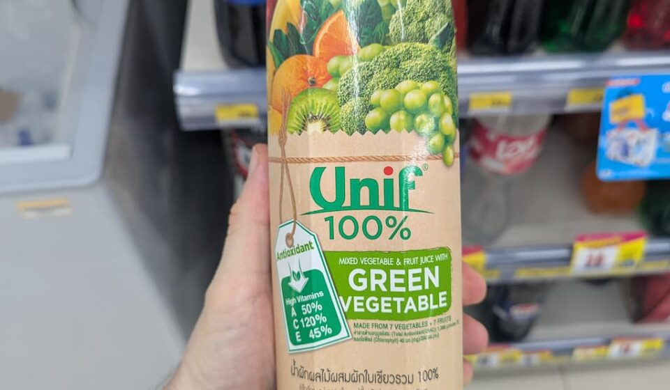 A hand holding a beige paper carton brik of Unif 100% Mixed Vegetable & Fruit Juice with Green Vegetable inside a 7-Eleven store in Thailand.