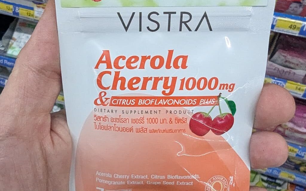 A hand holding a white and orange metallic zip-pouch of Vistra Acerola Cherry 1000mg dietary supplement tablets inside a store in Thailand.