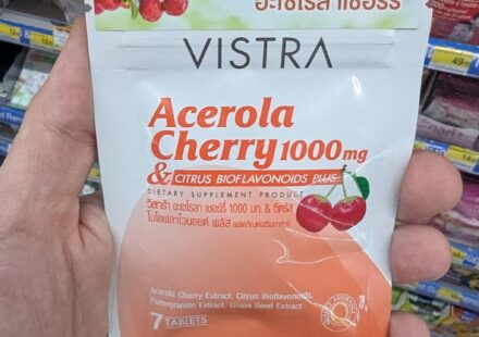 A hand holding a white and orange metallic zip-pouch of Vistra Acerola Cherry 1000mg dietary supplement tablets inside a store in Thailand.