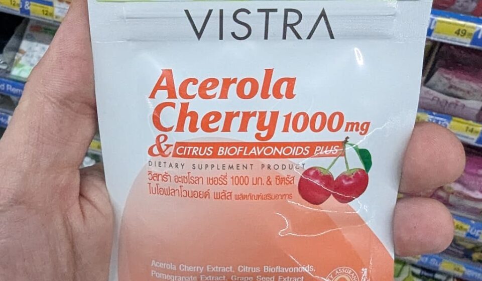 Vistra Acerola Cherry 1000mg: High-Dose Immune Defense in a Travel Pack A hand holding a white and orange metallic zip-pouch of Vistra Acerola Cherry 1000mg dietary supplement tablets inside a store in Thailand.