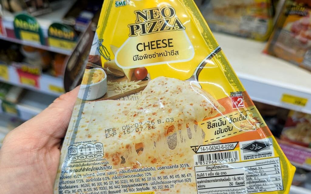A hand holding a yellow and white triangular packaging sleeve of an Alfredo Neo Pizza Cheese slice inside a 7-Eleven store in Thailand.