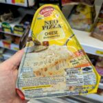A hand holding a yellow and white triangular packaging sleeve of an Alfredo Neo Pizza Cheese slice inside a 7-Eleven store in Thailand.