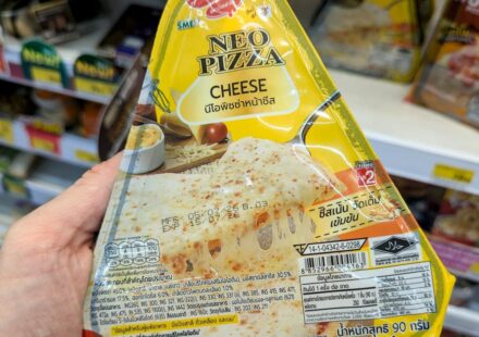 A hand holding a yellow and white triangular packaging sleeve of an Alfredo Neo Pizza Cheese slice inside a 7-Eleven store in Thailand.