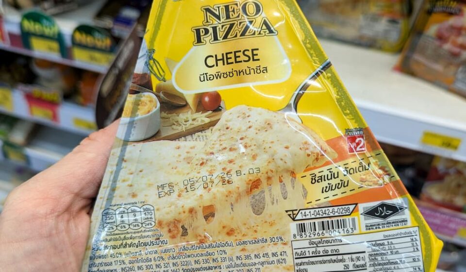 Alfredo Neo Pizza Cheese: A Flawed, Doughy Slice Designed for Immediate Stomach Filling A hand holding a yellow and white triangular packaging sleeve of an Alfredo Neo Pizza Cheese slice inside a 7-Eleven store in Thailand.