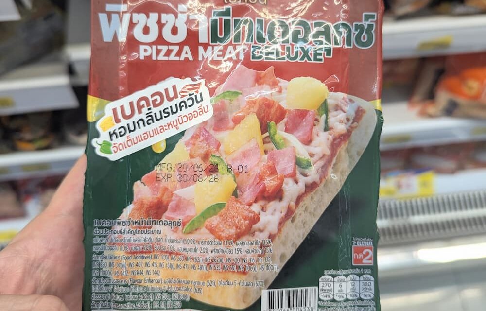 A hand holding a dark green and red packaging pouch of a Bakon Pizza Meat Deluxe individual slice inside a store in Thailand.
