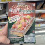 A hand holding a dark green and red packaging pouch of a Bakon Pizza Meat Deluxe individual slice inside a store in Thailand.