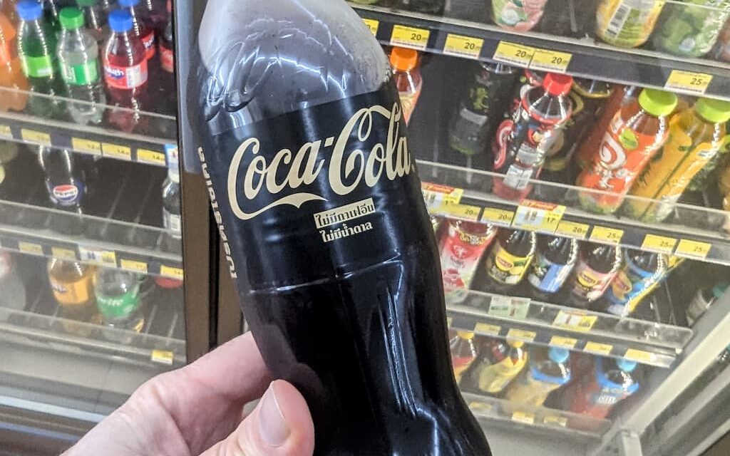 A hand holding a minimalist black bottle of Coca-Cola Zero Sugar Zero Caffeine with gold lettering inside a 7-Eleven store in Thailand.
