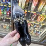 A hand holding a minimalist black bottle of Coca-Cola Zero Sugar Zero Caffeine with gold lettering inside a 7-Eleven store in Thailand.