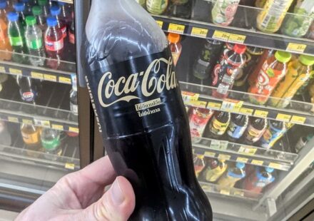 Coca-Cola Zero Sugar Zero Caffeine: The Minimalist Black Bottle That Strips Away Everything A hand holding a minimalist black bottle of Coca-Cola Zero Sugar Zero Caffeine with gold lettering inside a 7-Eleven store in Thailand.