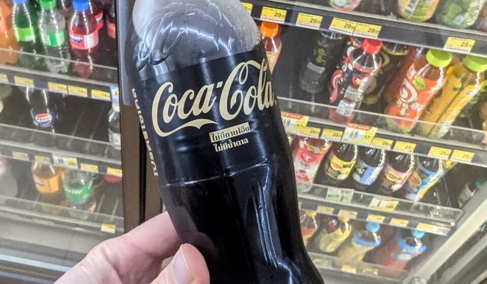 A hand holding a minimalist black bottle of Coca-Cola Zero Sugar Zero Caffeine with gold lettering inside a 7-Eleven store in Thailand.