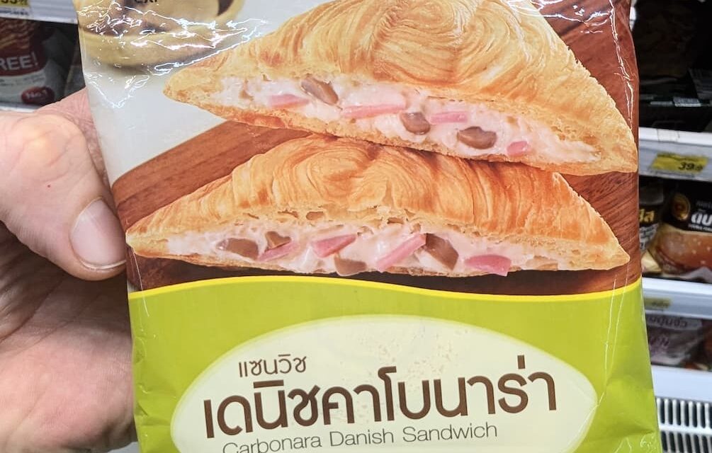 A hand holding a light green and white plastic pouch packaging of an Ezygo Carbonara Danish Sandwich in front of store display shelves in Thailand.