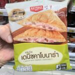 Chester’s Grilled Hot and Spicy Chicken: A Savory Classic That Demands Maximum Heat A hand holding a light green and white plastic pouch packaging of an Ezygo Carbonara Danish Sandwich in front of store display shelves in Thailand.