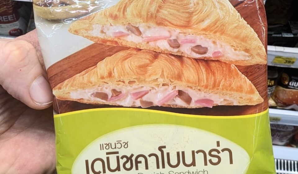 A hand holding a light green and white plastic pouch packaging of an Ezygo Carbonara Danish Sandwich in front of store display shelves in Thailand.