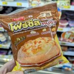 A hand holding a brown plastic bag of an Ezygo Four Cheese Burger with Pork and Cheese in front of refrigerator shelves at a store in Thailand.