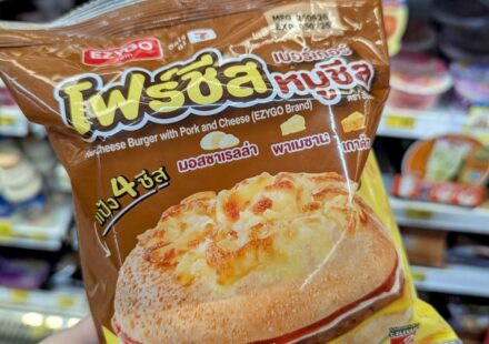 A hand holding a brown plastic bag of an Ezygo Four Cheese Burger with Pork and Cheese in front of refrigerator shelves at a store in Thailand.
