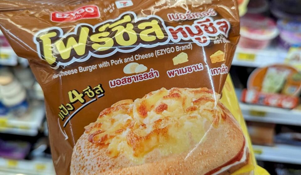 A hand holding a brown plastic bag of an Ezygo Four Cheese Burger with Pork and Cheese in front of refrigerator shelves at a store in Thailand.