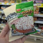 A hand holding a cream and green plastic pouch packaging of an Ezygo Grilled Pork Quinoa Sticky Rice Burger inside a store in Thailand.