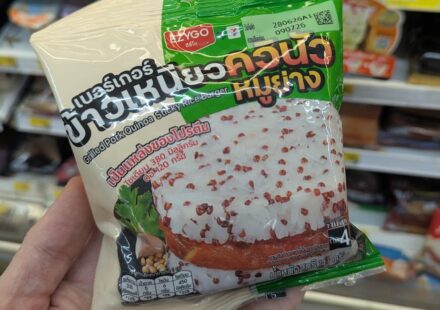 Ezygo Grilled Pork Quinoa Sticky Rice Burger: A Flavorful Street-Food Fusion with Serious Heat A hand holding a cream and green plastic pouch packaging of an Ezygo Grilled Pork Quinoa Sticky Rice Burger inside a store in Thailand.